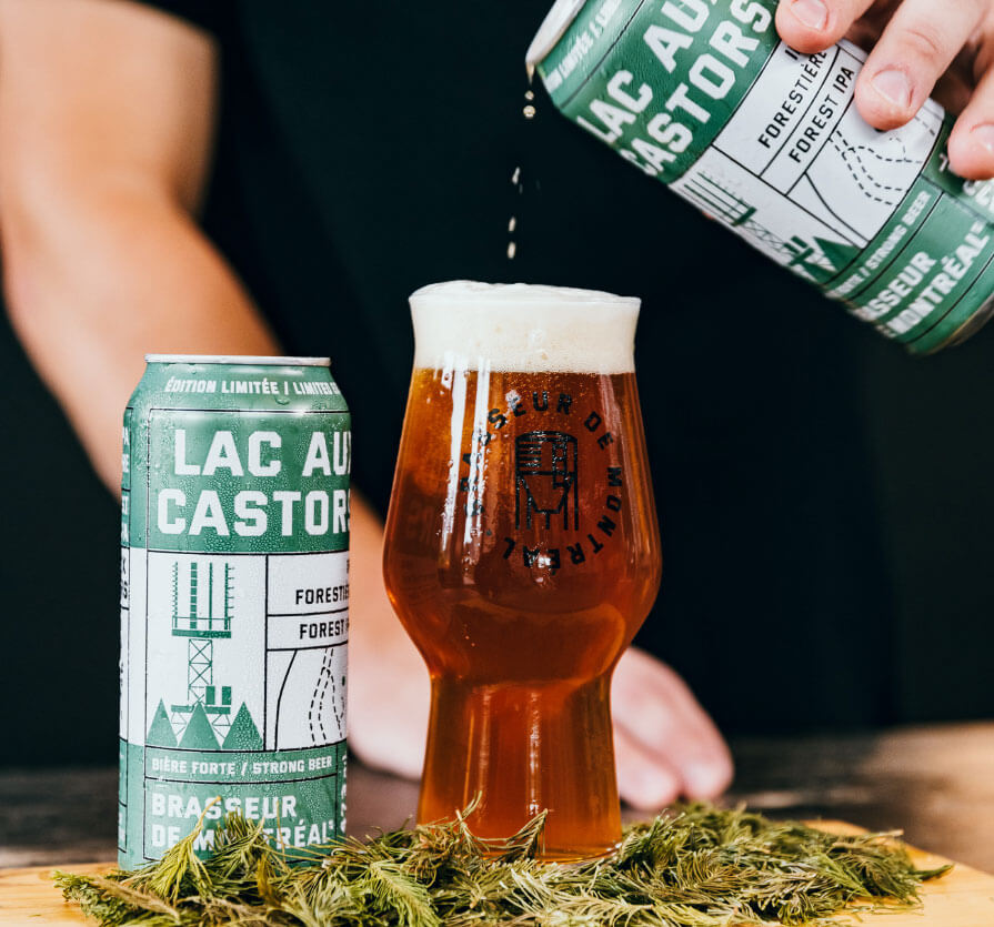 Glass of amber beer with foam, surrounded by green sprigs, next to two green cans labeled “Lac aux Castors IPA.” One can is pouring beer into the glass.